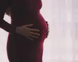 Kobieta w ciąży w bordowej sukience delikatnie obejmuje dłońmi brzuszek.