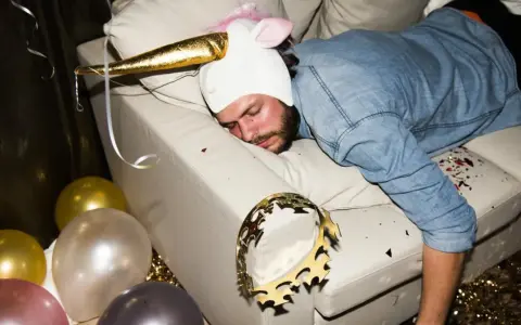 Mężczyzna w stroju jednorożca śpi na kanapie po imprezie. Balony i konfetti sugerują, że to może być sposób na to, jak pozbyć się kaca.