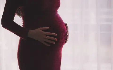 Kobieta w ciąży w bordowej sukience delikatnie obejmuje dłońmi brzuszek.