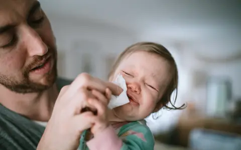 Ojciec delikatnie wyciera nosek niemowlaka, który ma katar i kaszel, ale nie gorączkę. Dziecko płacze, czując się źle.