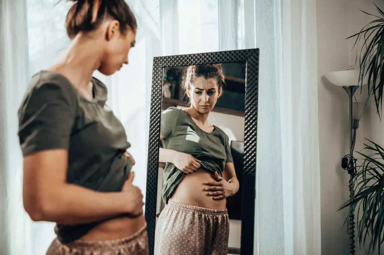 Kobieta w piżamie dotyka brzucha, patrząc w lustro z niepokojem. Czy przy menopauzie boli brzuch?
